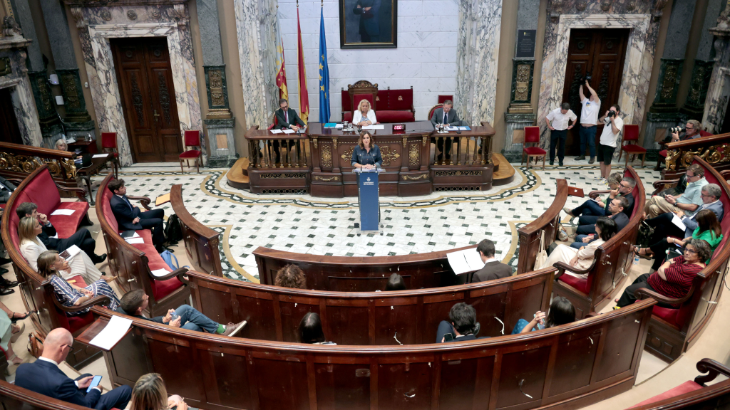 a woman standing at a podium with a group of people sitting in the background